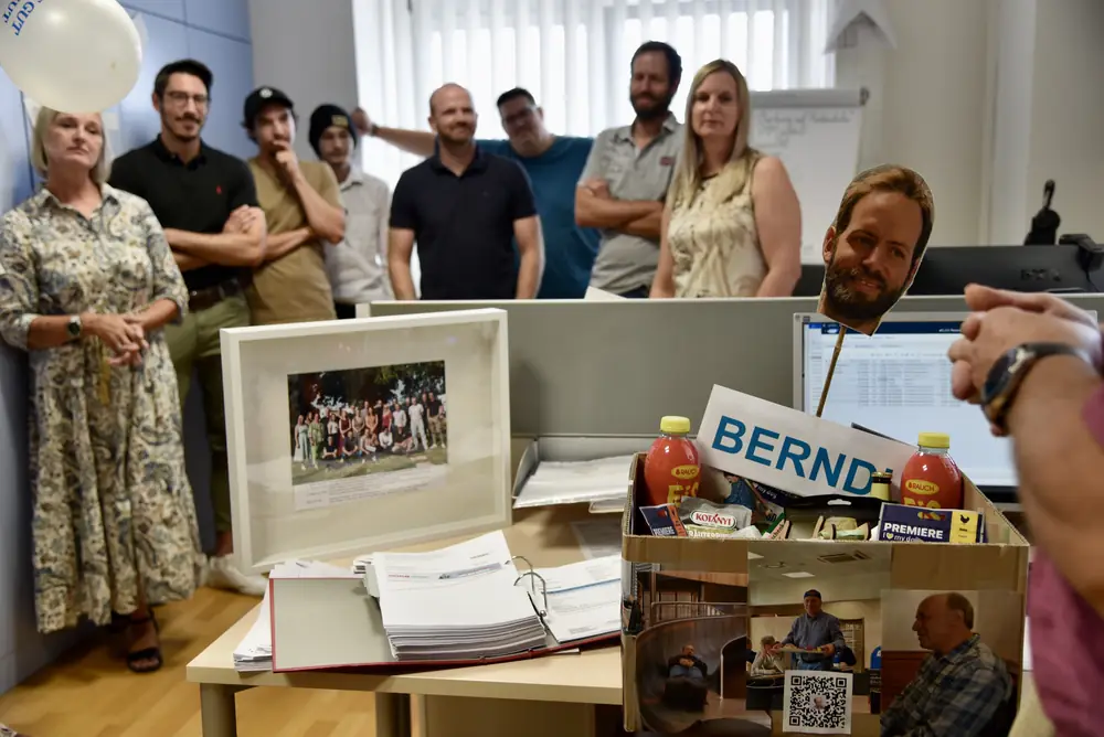 Das gerahmte Gruppenfoto und die Geschenkkiste auf dem Schreibtisch, das Team im Hintergrund
