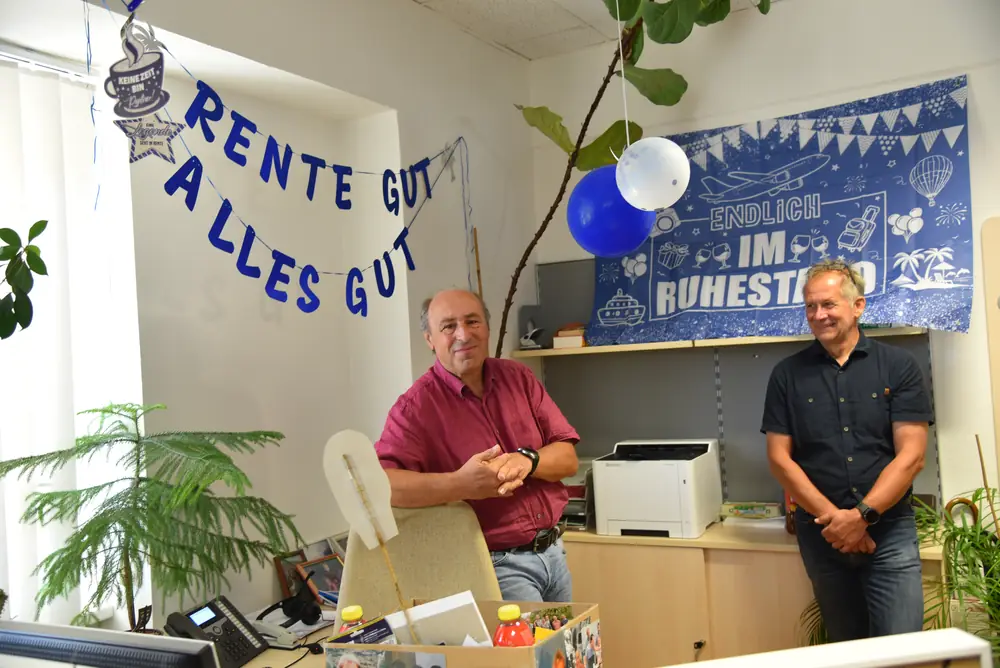 Günter und ein Kollege lachen vor der „Rente gut, alles gut“-Girlande